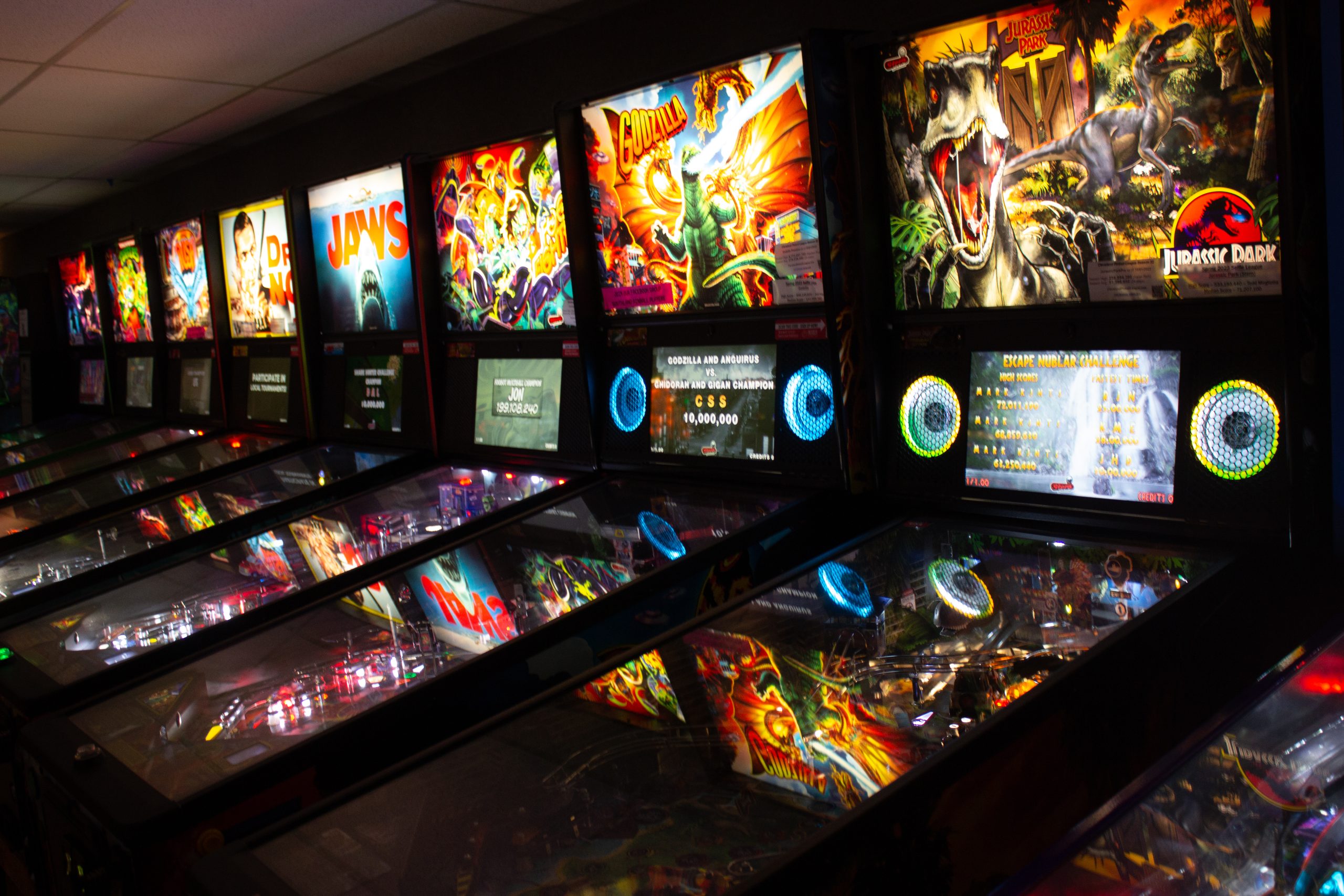 A row of brightly lit pinball machines in a dark room.